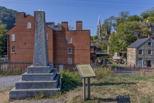 John Brown Monument; with John Browns Fort behind it and St Peters Roman Catholic Church in the distance.   Sign Reads: Commemorated here is the original location of the ‘John Brown Fort’ – the Federal Armory’s fire-engine house, where abolitionist John Brown and his raiders were captured by US Marines on October 18, 1859.  If you look to the south you will see the Fort almost 150 feet from here.  The Fort was first moved in 1892 and its original foundation covered by the railroad in 1892.